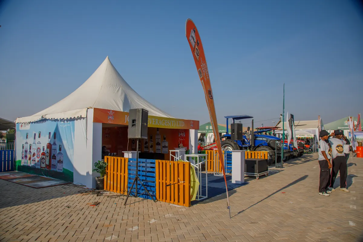 John Beverages (JBL) branded tent along the exhibitor row at Nane Nane Grounds, Dodoma, with agricultural machinery and neighbouring stands in the background